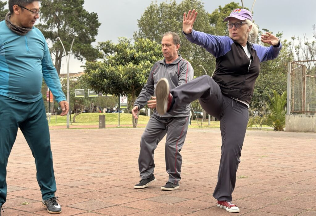a woman practicing a tai chi kick while two men observe and practice